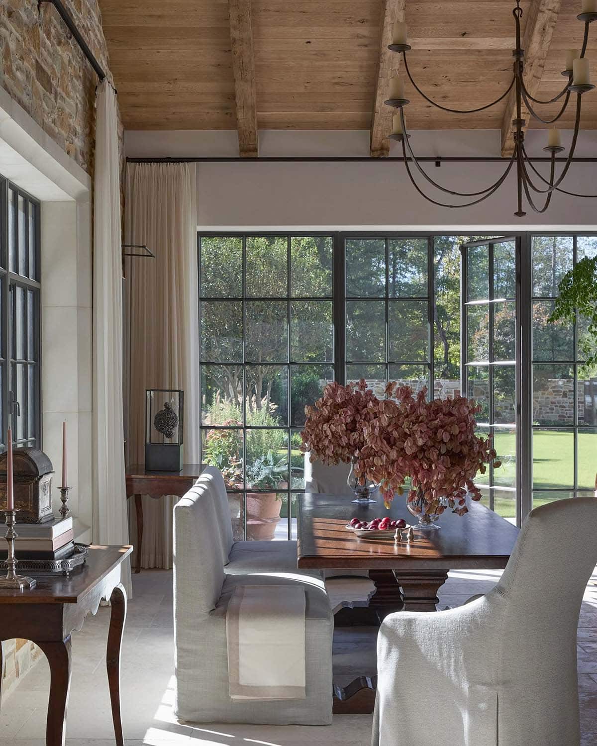 Dining room with slipcovered chairs, wood trestle table, dried floral arrangement, and steel garden doors
