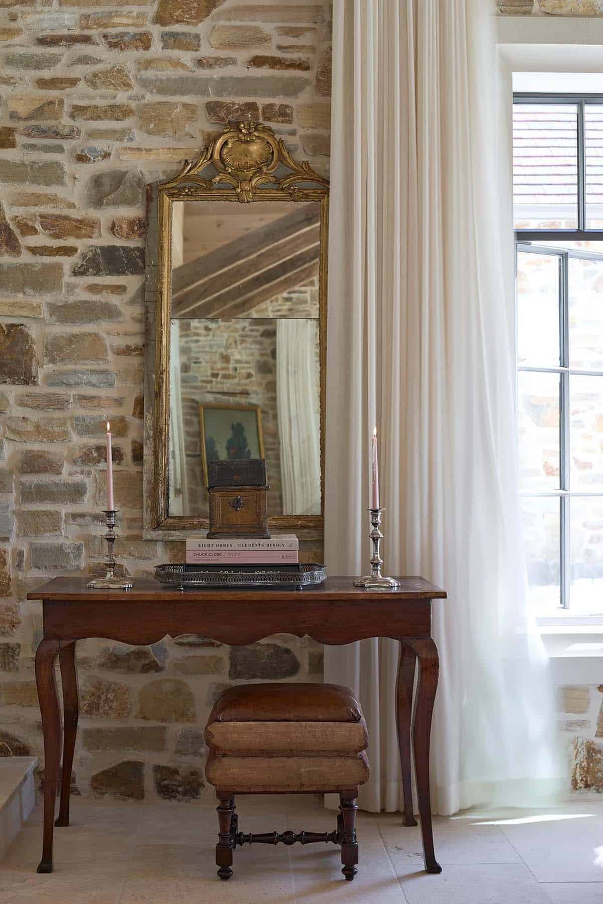 Antique console table with gilt mirror, silver candlesticks, and leather stool against stone wall