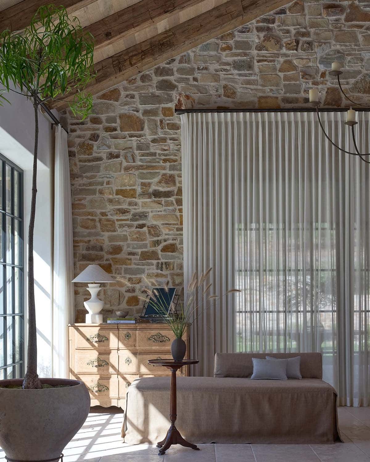 Living room corner with linen daybed, antique dresser, sheer curtains, and stone wall