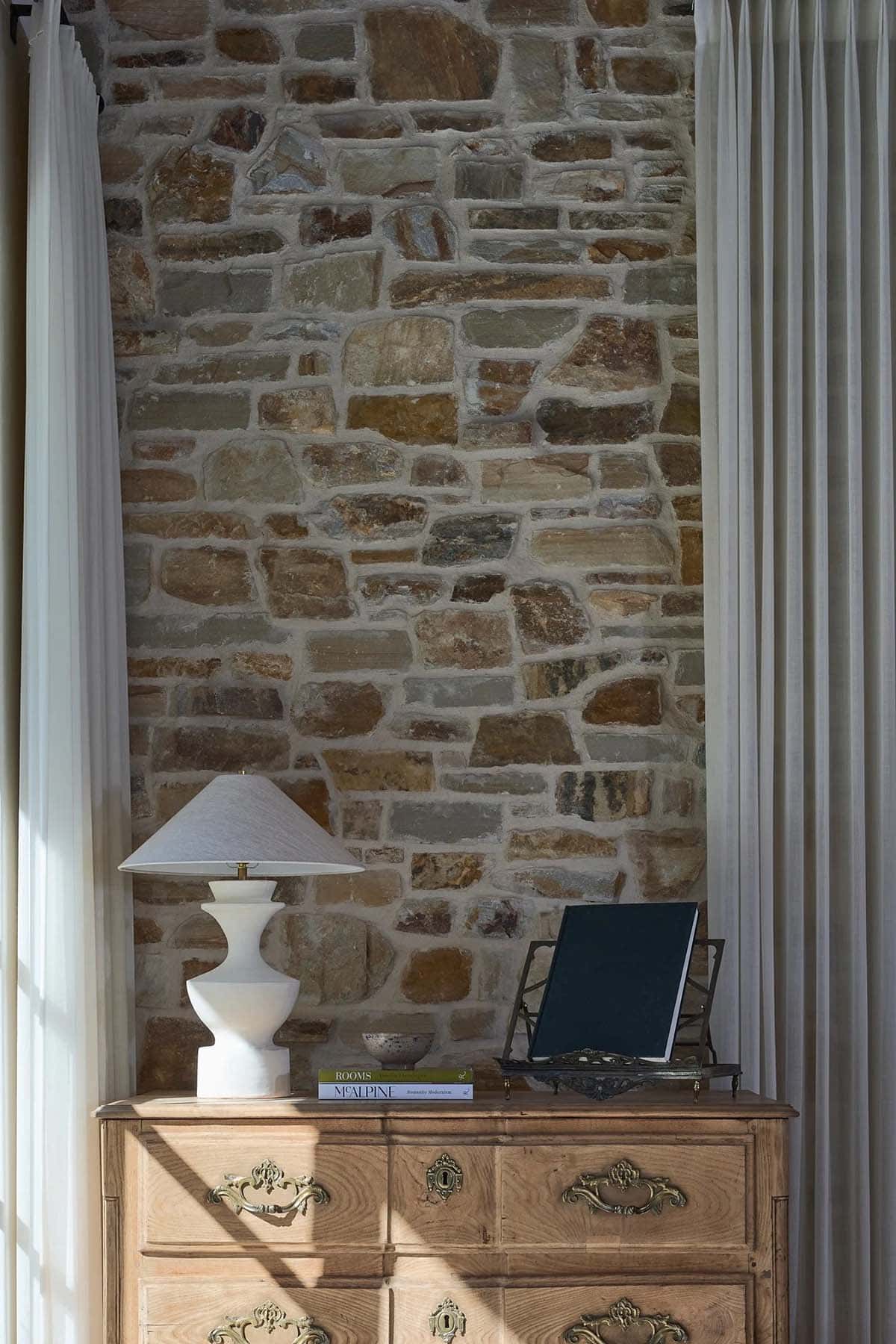 Antique dresser detail with ceramic lamp, design books, and exposed stone wall