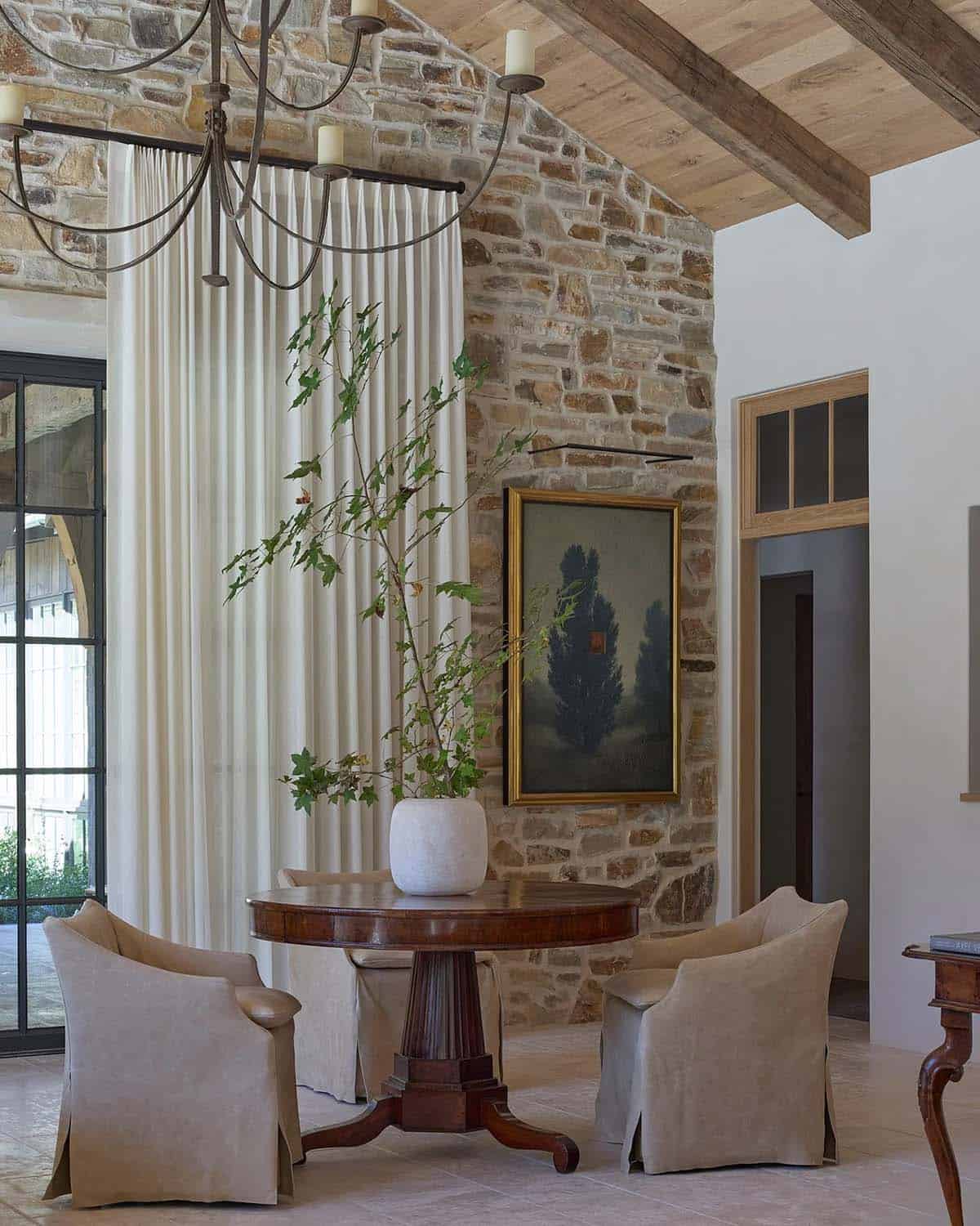 Sitting area with round pedestal table, linen chairs, tall curtains, and landscape painting on stone wall