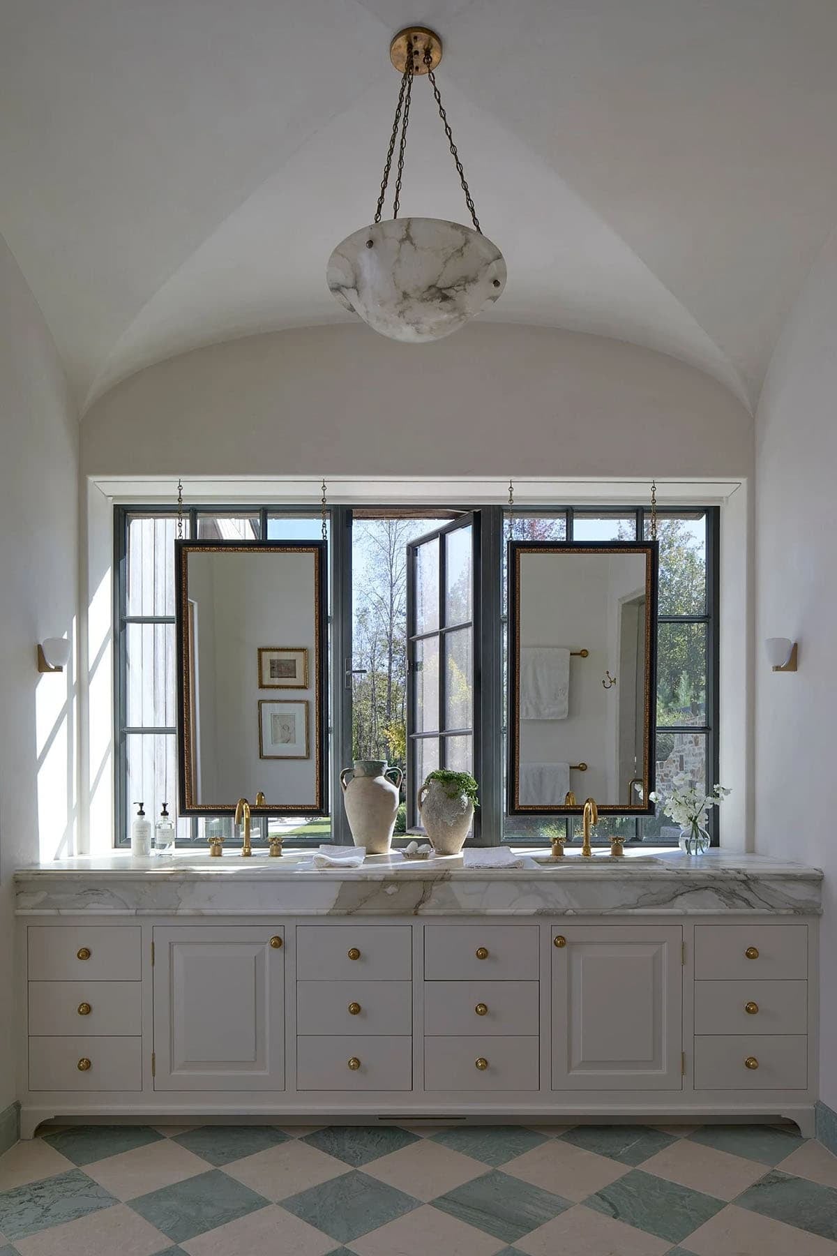 Primary bathroom double vanity with marble countertop, brass fixtures, alabaster pendant, and checkerboard marble floor