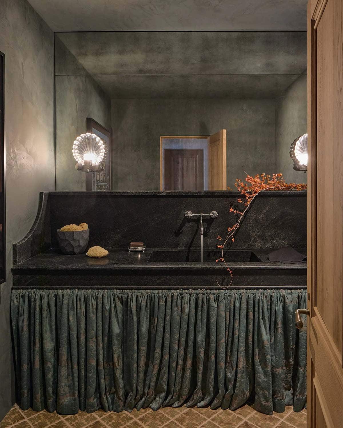 Powder room with dark plaster walls, black stone sink, skirted vanity, and shell-shaped sconces