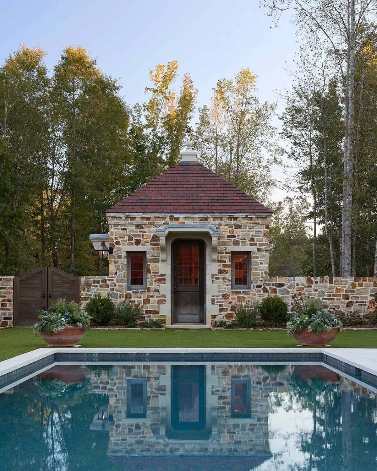 Stone pool house with pyramid roof reflected in still rectangular pool at dusk
