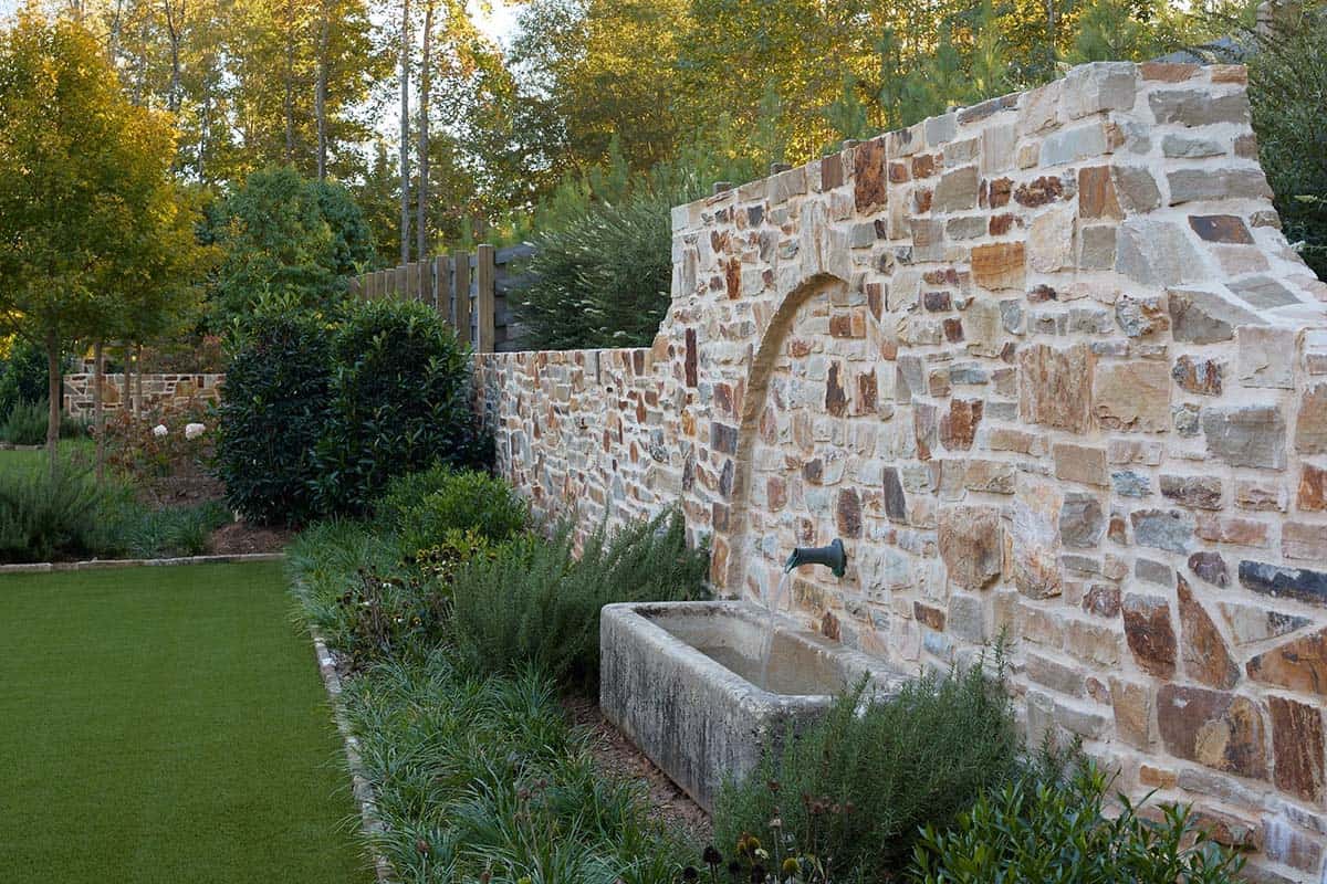 Stone garden wall with arched niche, antique stone trough fountain, and perennial border