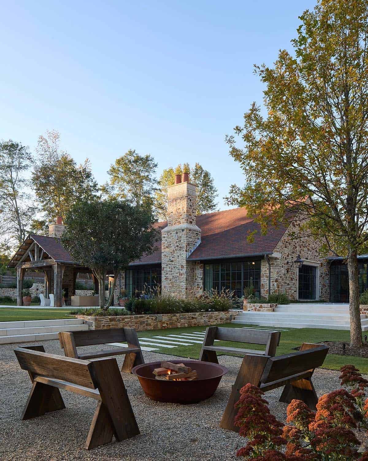 Fire pit seating area with wood benches, steel fire bowl, and stone home facade beyond at dusk
