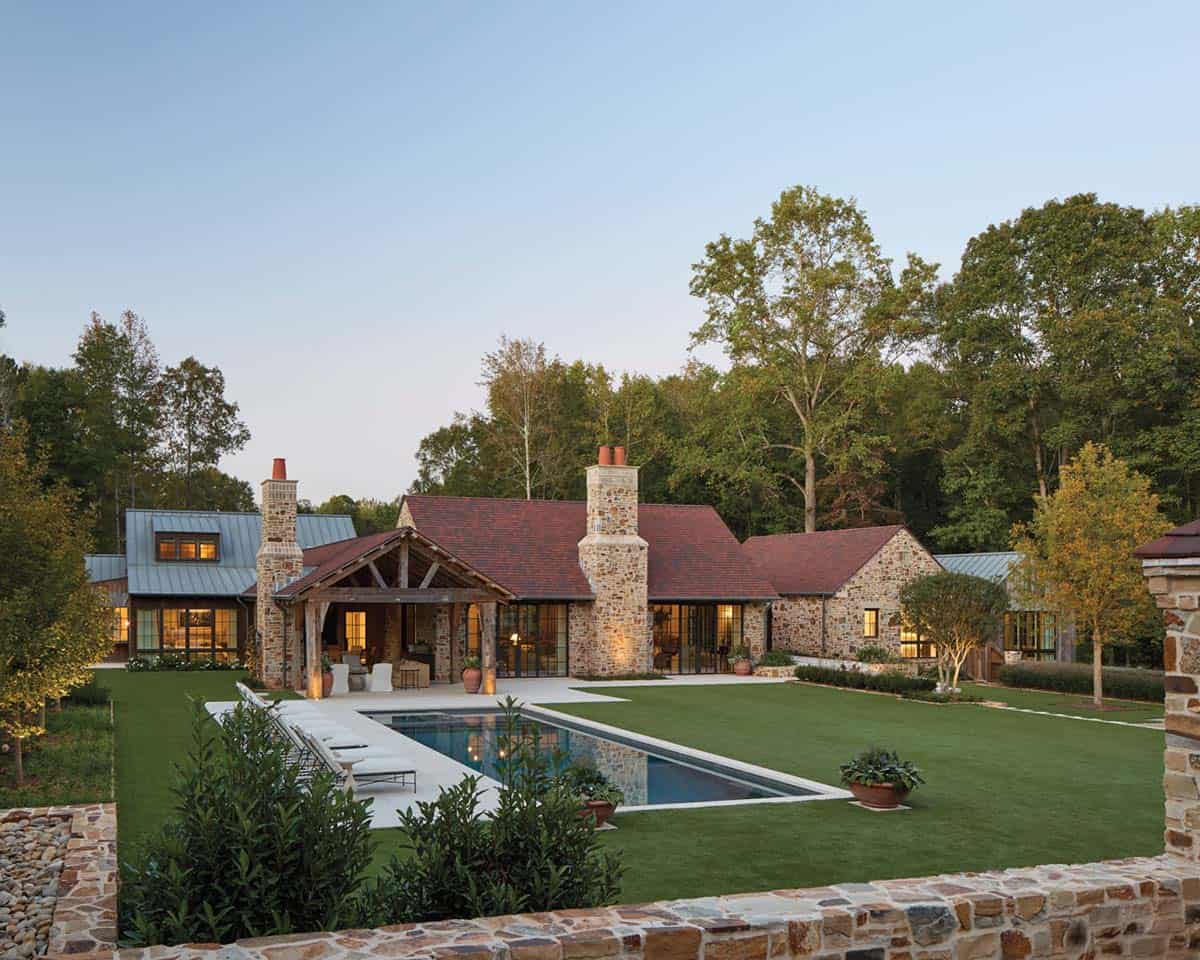 Elegant stone home in Serenbe Georgia with red roof, twin chimneys, timber pergola, and rectangular pool at dusk