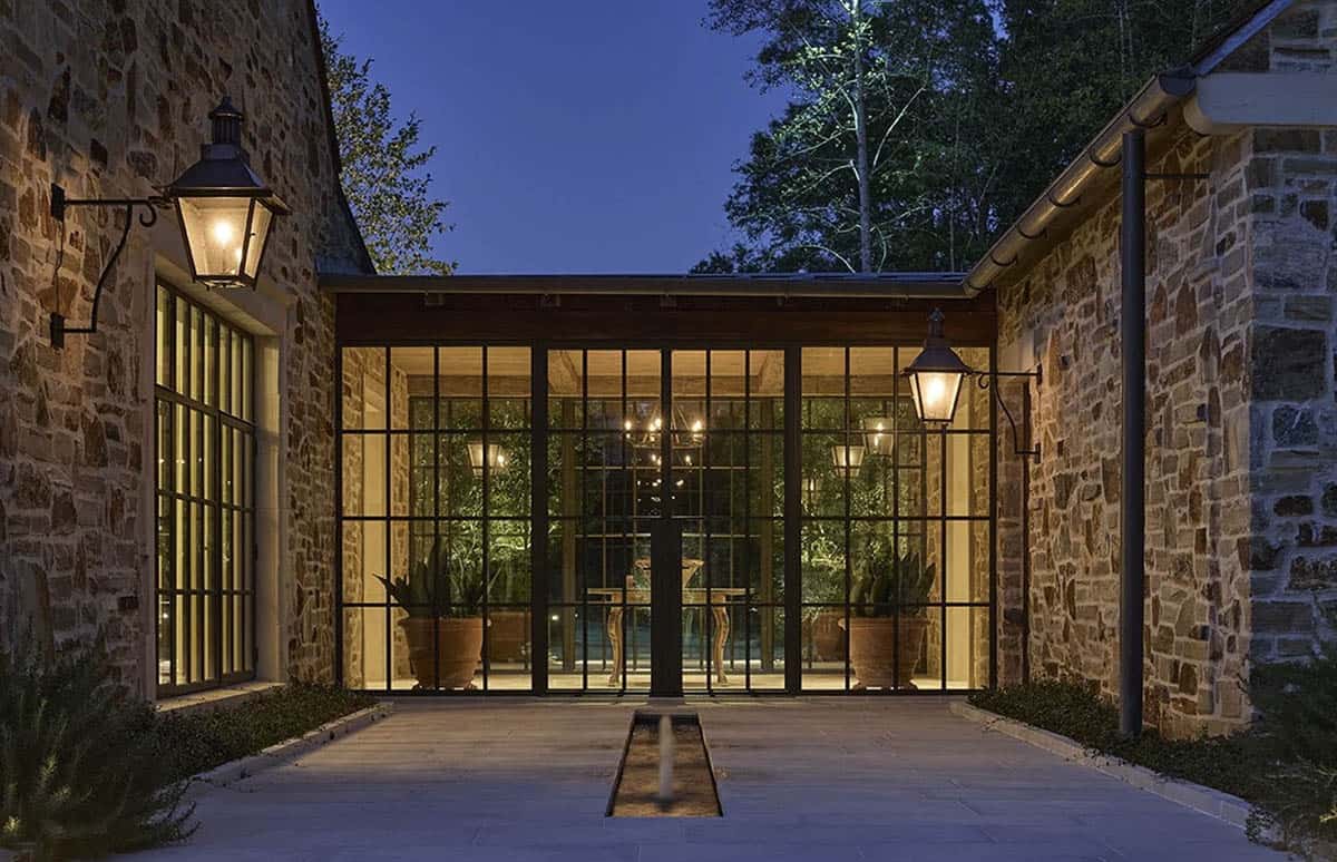 Stone courtyard at dusk with steel-framed glazed entrance, copper gas lanterns, and narrow water feature