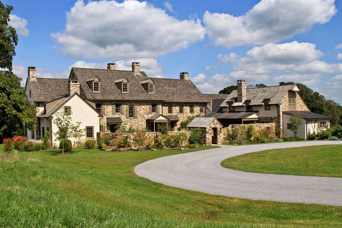 Expansive fieldstone manor house with cedar shake roof and white clapboard wing viewed from sweeping driveway