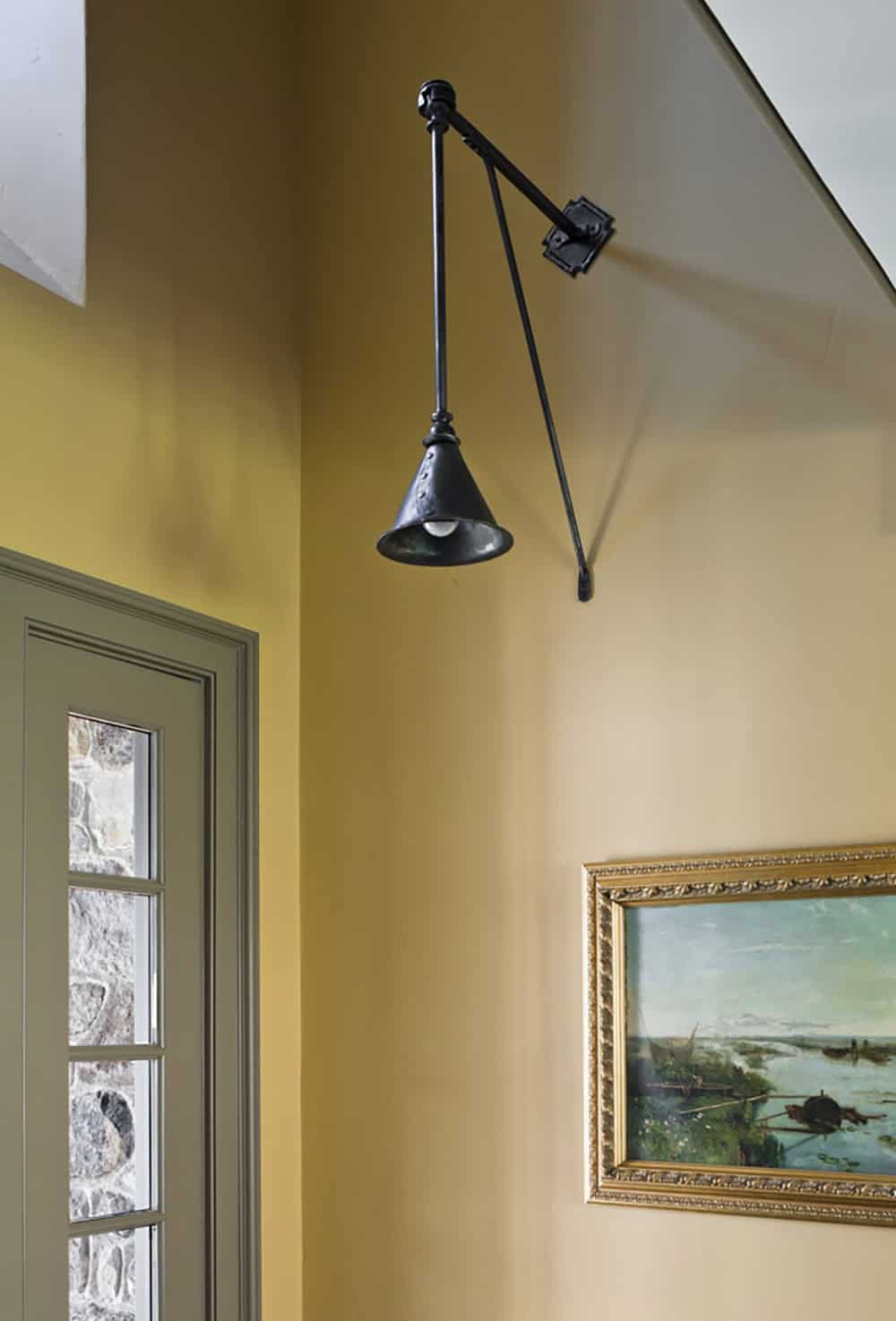 Black wrought-iron adjustable wall sconce against mustard yellow wall with landscape painting below