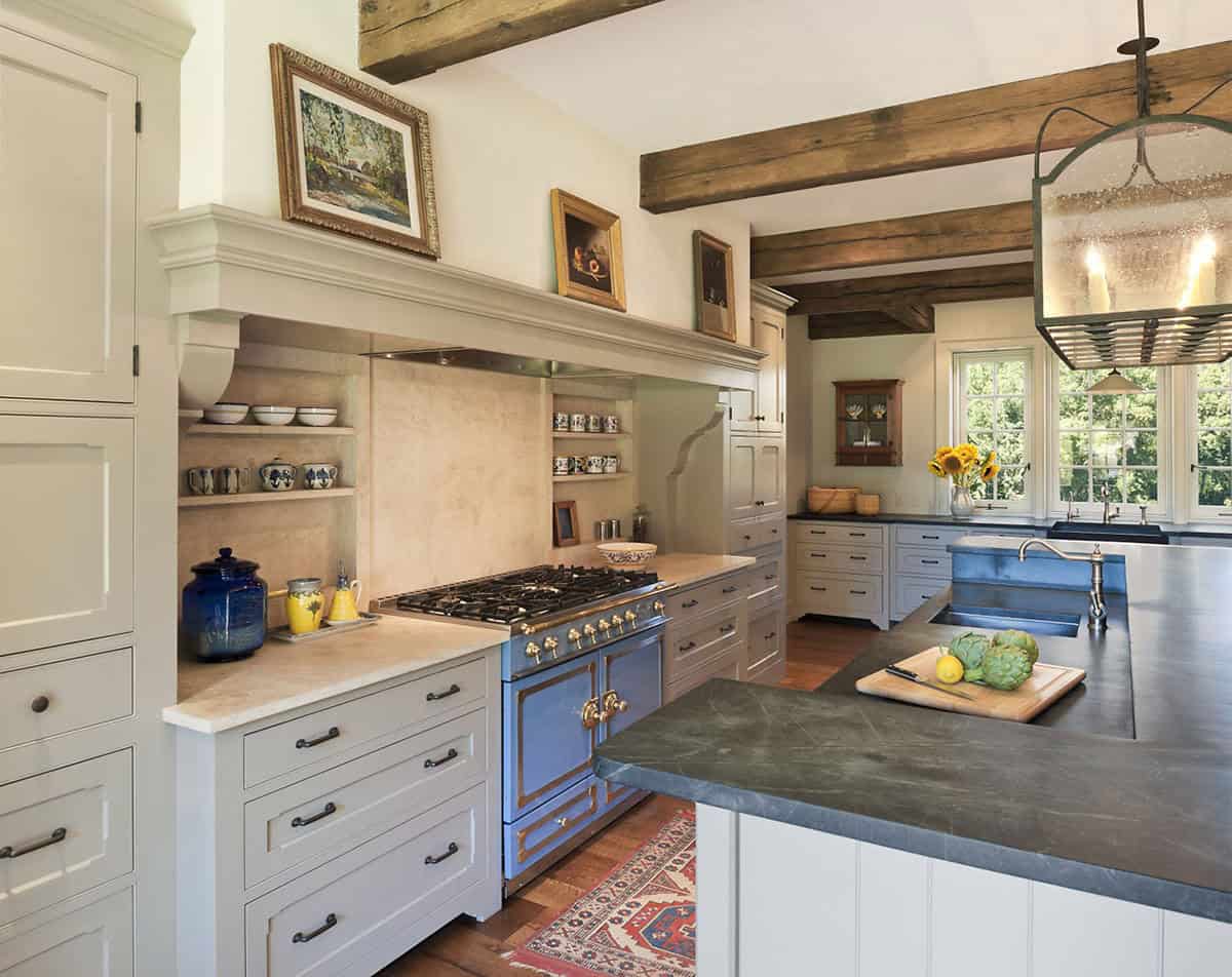 Farmhouse kitchen showing blue La Cornue range, soapstone island, and stone backsplash under timber beams