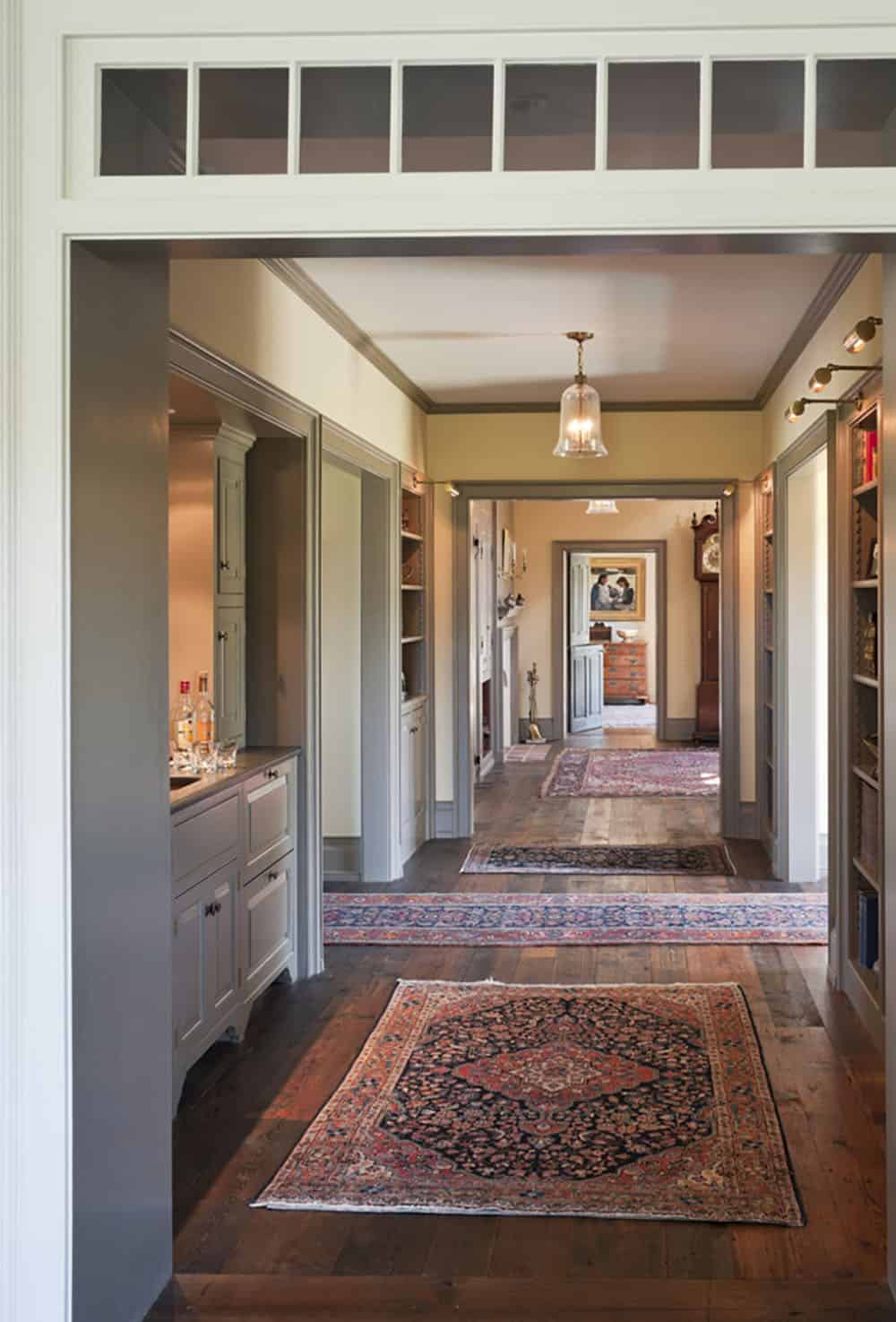 Long interior hallway with wide-plank floors, layered Persian rugs, and pendant lights leading through enfilade rooms