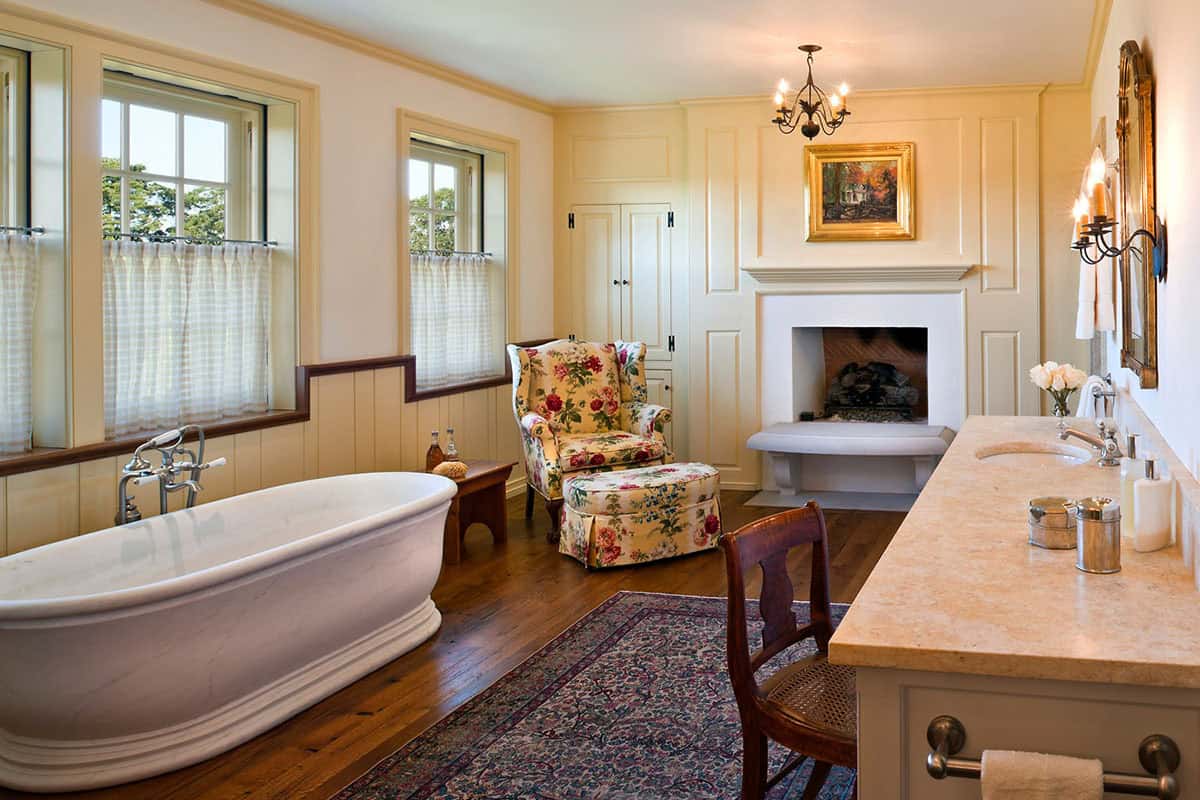 Main bathroom with freestanding marble soaking tub, fireplace, floral armchair, and wide-plank floors