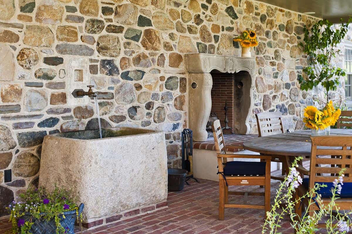 Covered stone porch with antique stone trough fountain, carved limestone fireplace surround, and teak dining furniture
