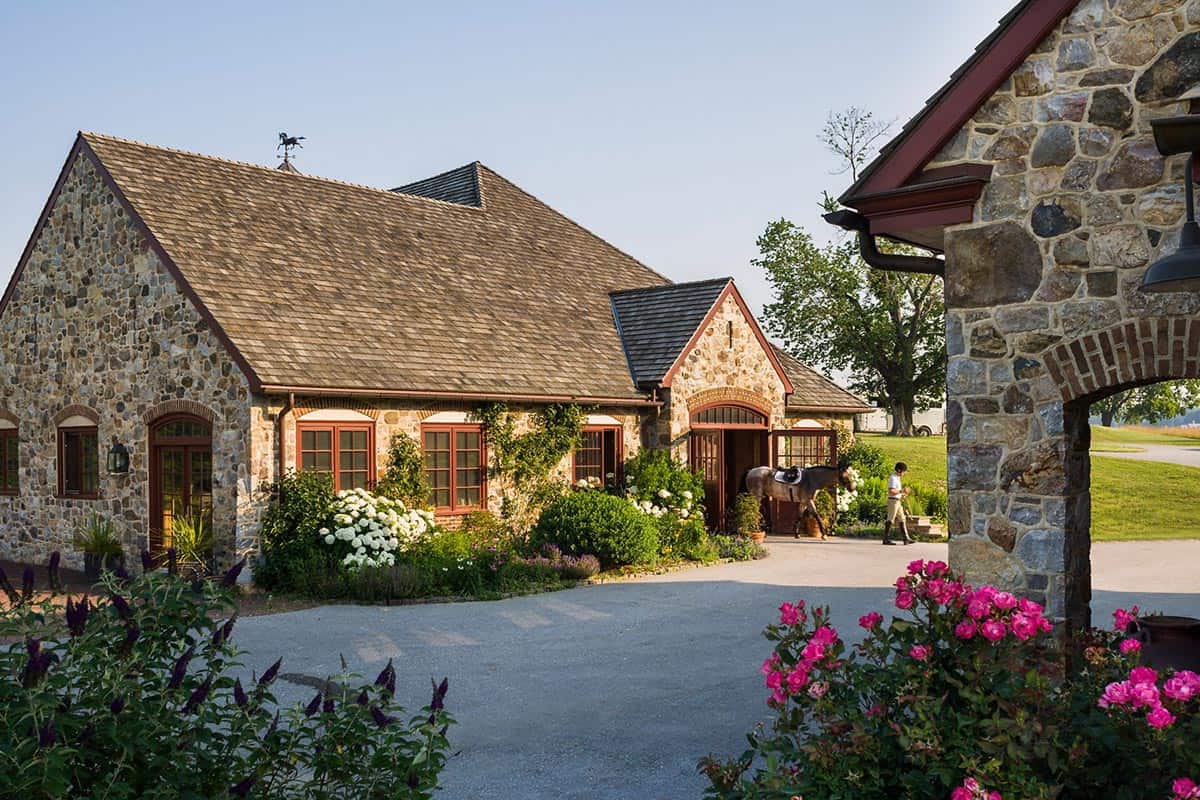 Fieldstone stable building with red trim, horse being led out, and flowering garden borders at dusk