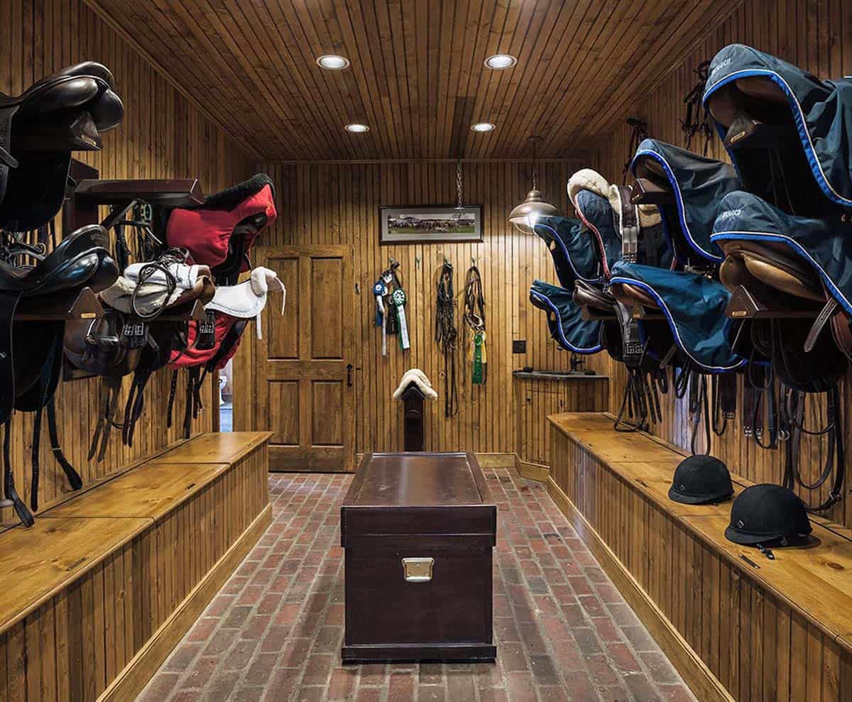 Wood-paneled tack room with saddles and horse blankets on racks, show ribbons, and brick floor