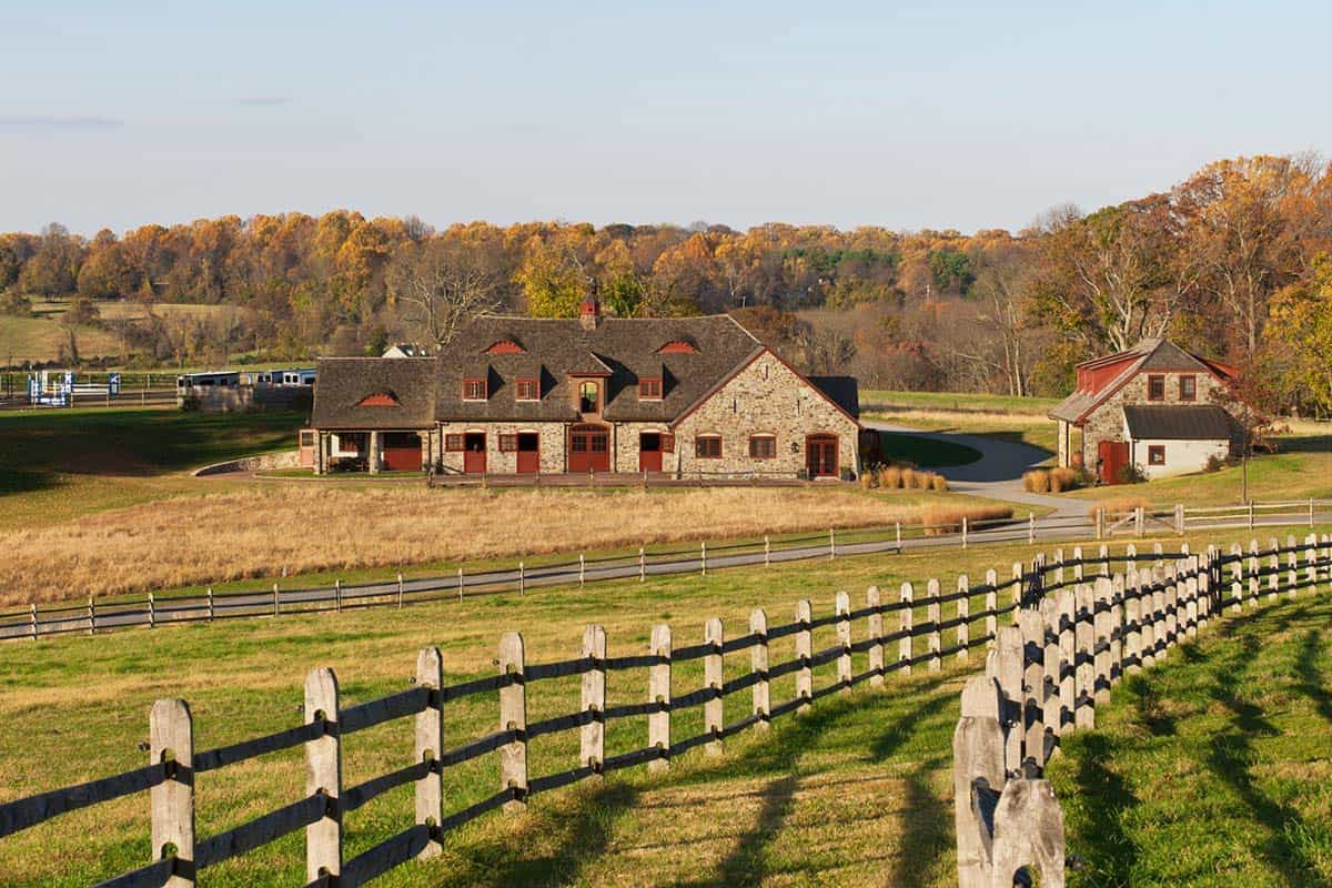 Aerial view of fieldstone equestrian barn complex with split-rail fencing and autumn foliage landscape
