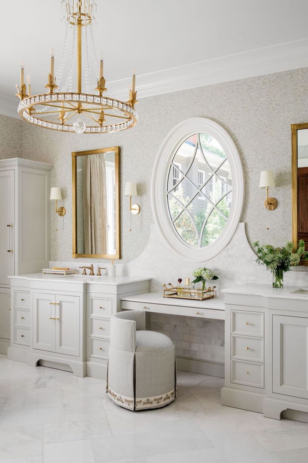 Primary bathroom vanity with dual sinks, oval leaded-glass window, and crystal chandelier