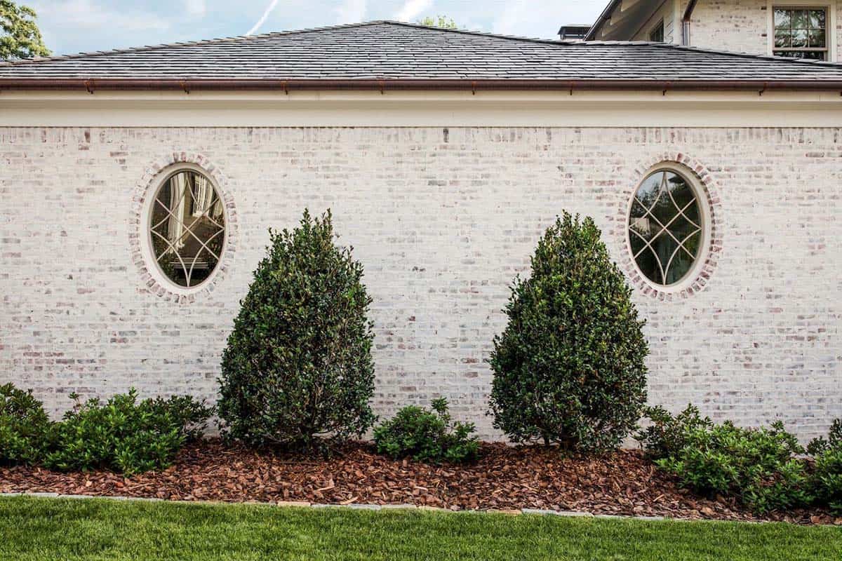 White painted brick exterior wall with two oval leaded-glass windows and manicured shrubs