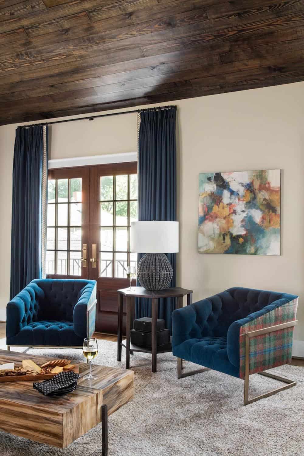 Carriage house sitting room with tufted blue velvet chairs, dark wood ceiling, and abstract art