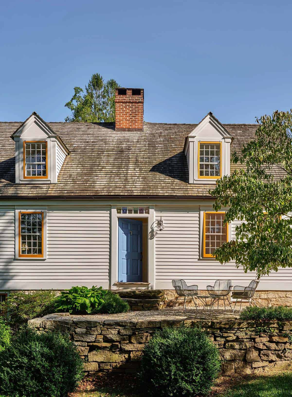 Historic colonial farmhouse exterior with blue front door, yellow window trim, dormer windows, and stone retaining wall