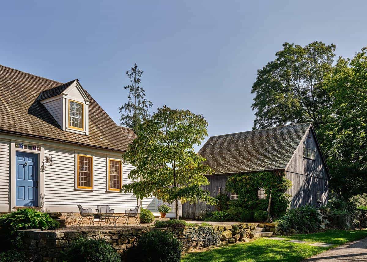 White clapboard colonial farmhouse with yellow-trimmed windows, blue door, and weathered barn outbuilding