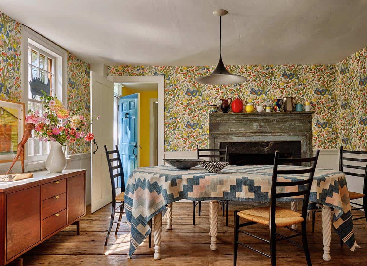 Colorful dining room with floor-to-ceiling botanical wallpaper, patchwork tablecloth, original fireplace, and black pendant light