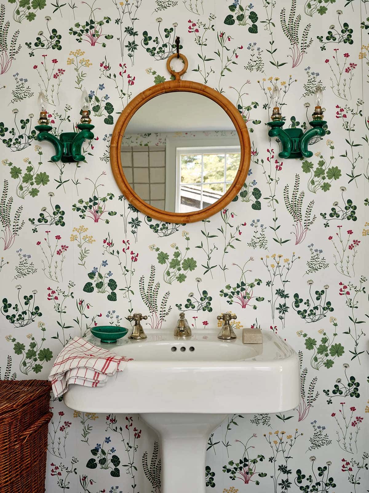 Powder room with botanical floral wallpaper, pedestal sink, rattan mirror, and green ceramic sconces