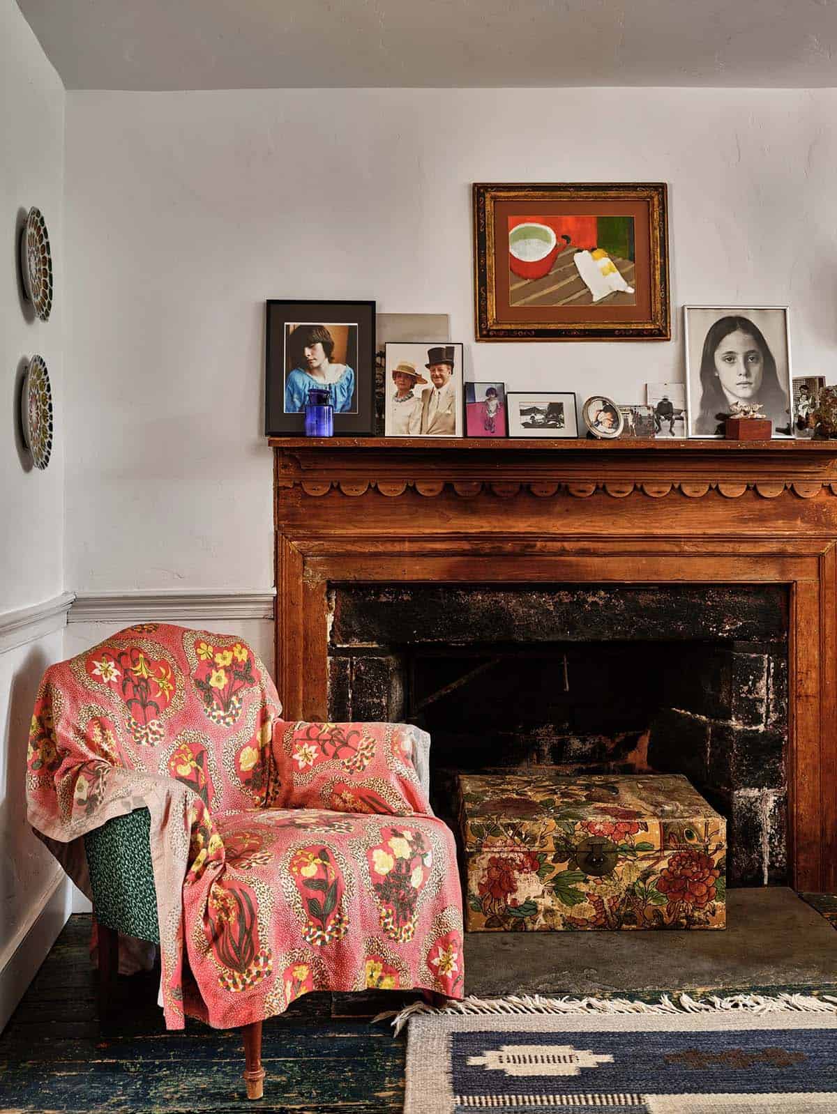 Bedroom fireplace nook with carved wood mantel, family photos, floral-draped armchair, and patterned footstool