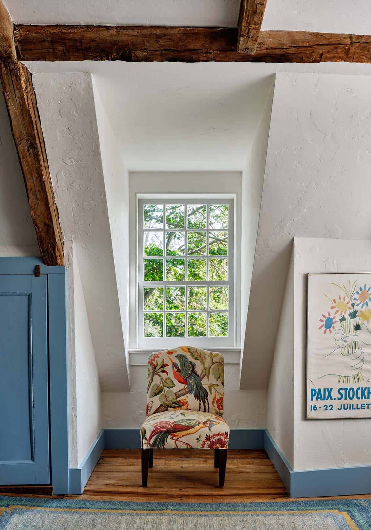 Attic landing nook with exposed timber beams, deep dormer window, floral accent chair, and blue painted armoire