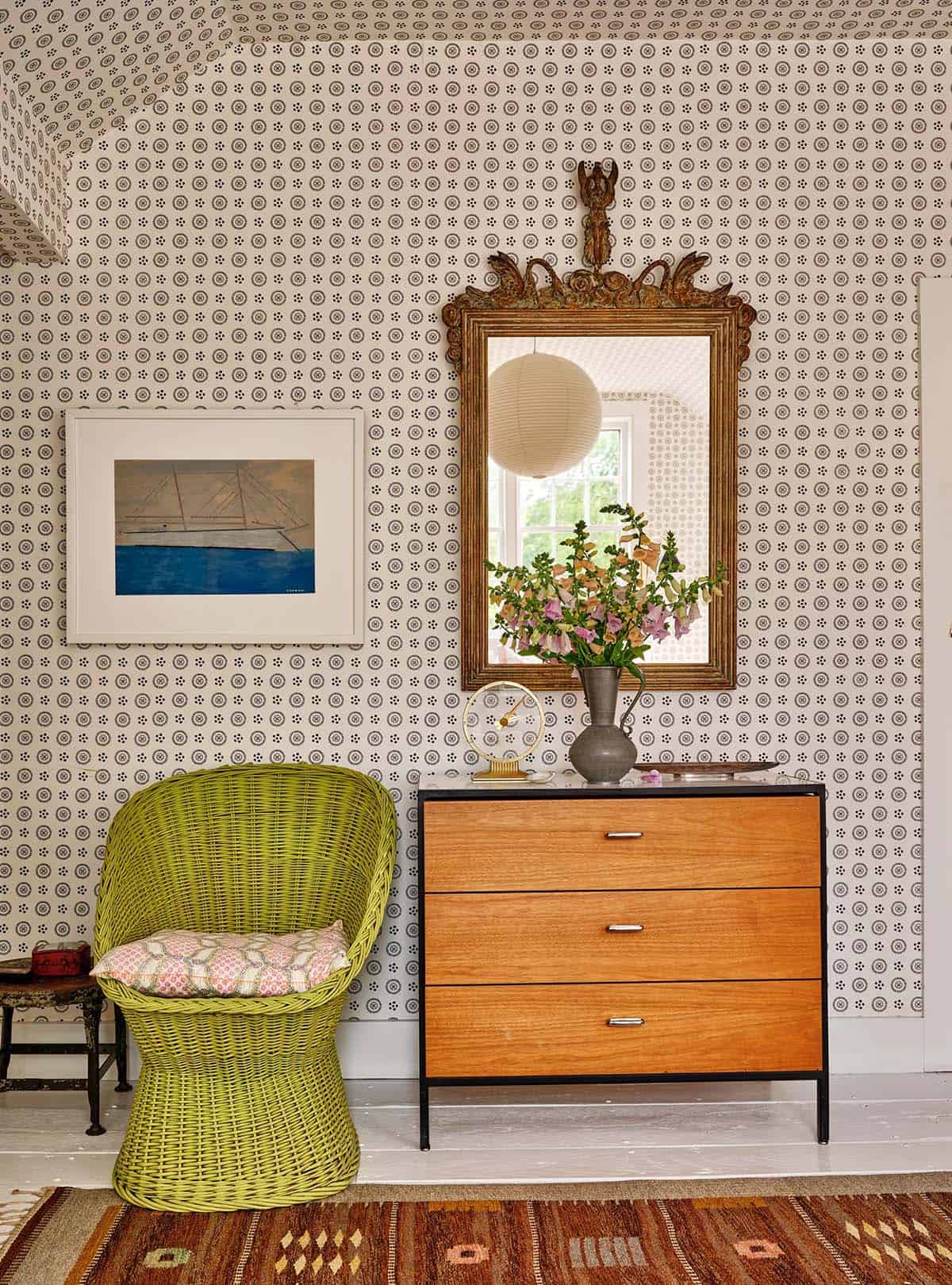 Bedroom corner with all-over geometric wallpaper, chartreuse wicker chair, mid-century dresser, and ornate gilt mirror