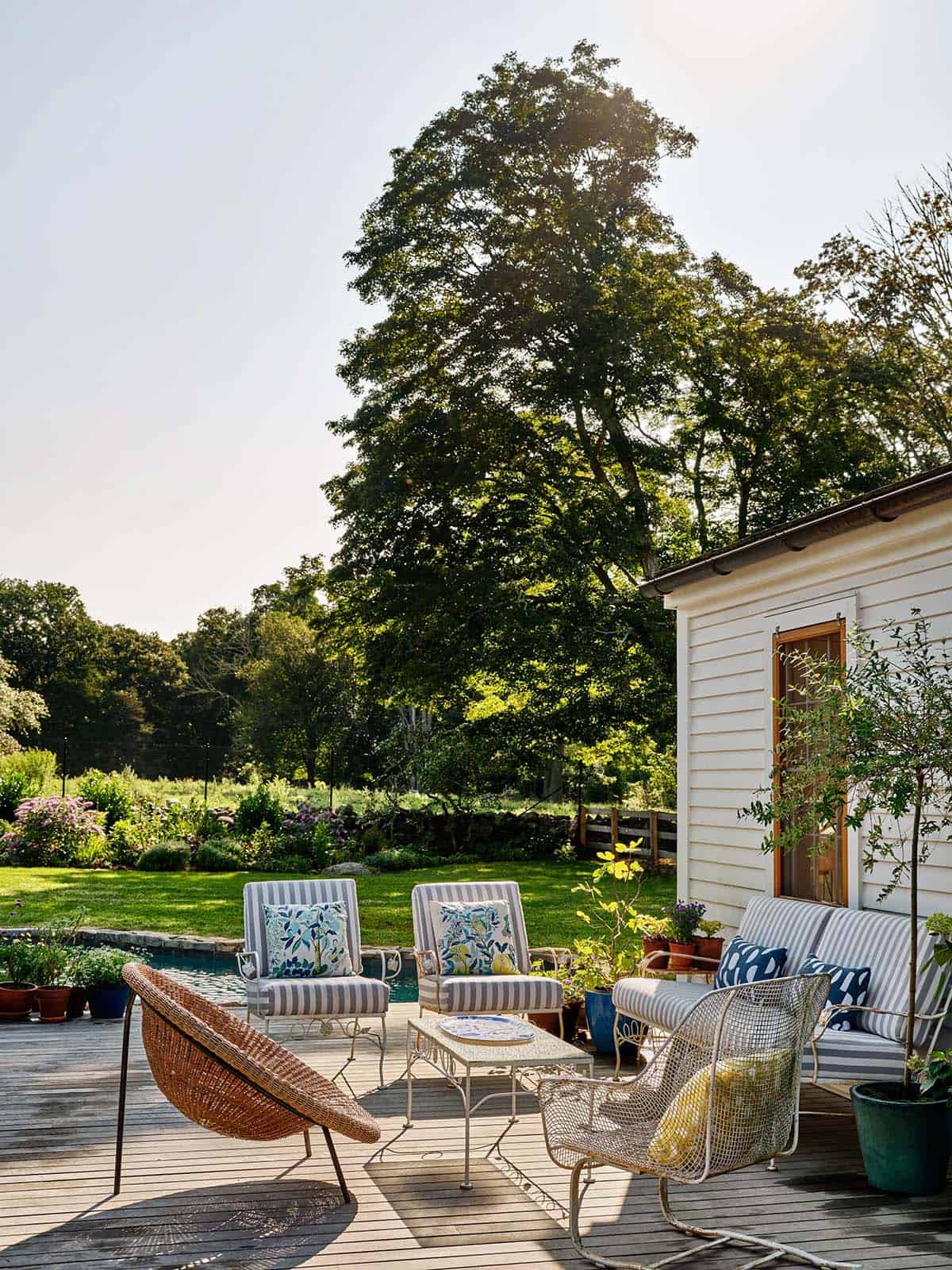 Sunny backyard deck with striped outdoor furniture, wicker lounge chair, potted plants, and garden views beyond