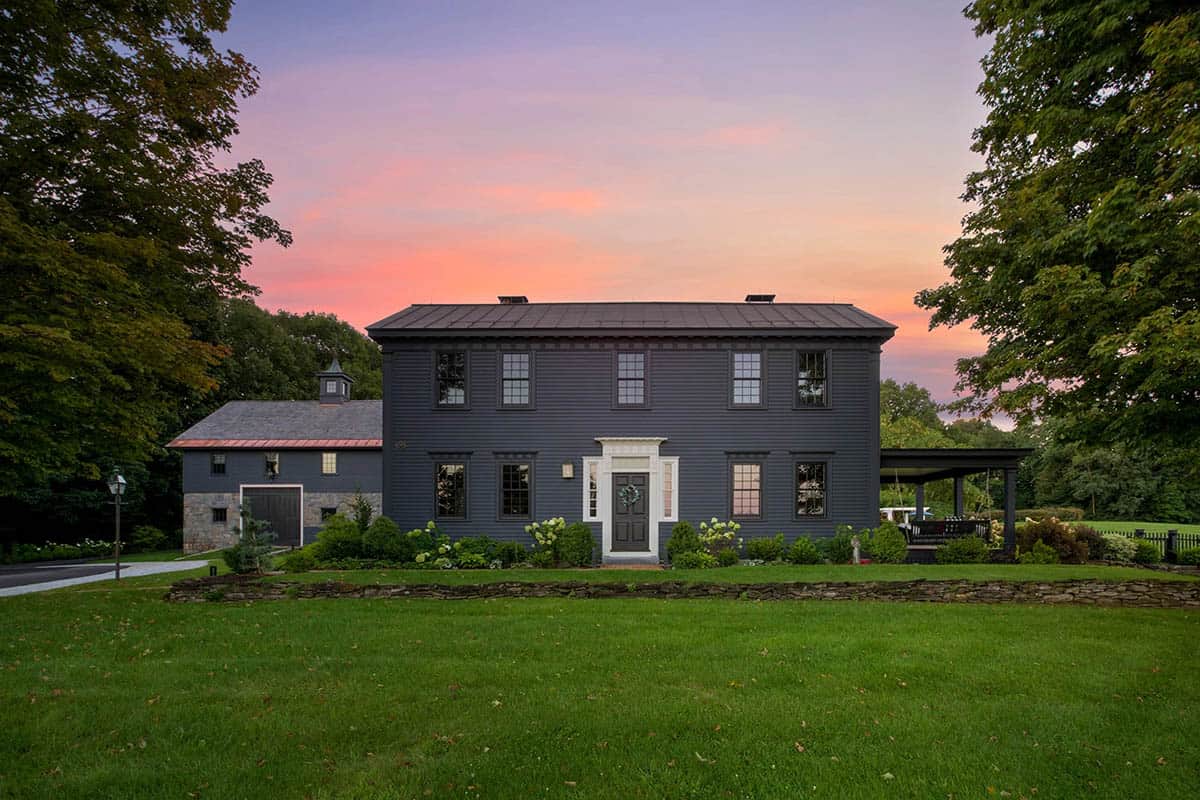 Restored colonial farmhouse with dark navy siding, white entry door, and attached barn at sunset