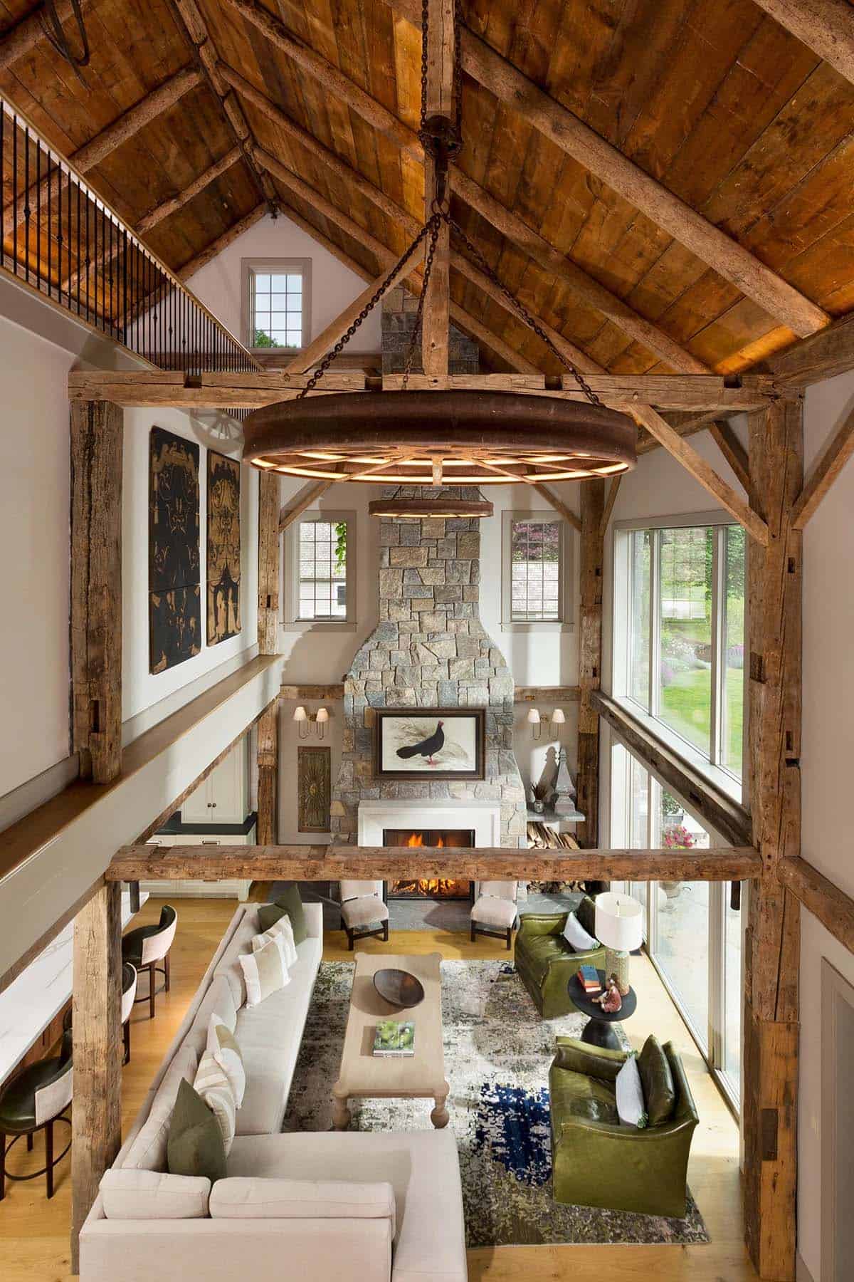 Double-height barn great room with original timber frame, wagon wheel chandelier, stone fireplace, and open loft above
