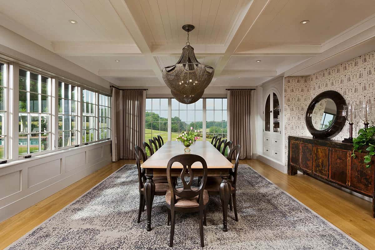 Dining room with coffered ceiling, ornate pendant chandelier, dark wood chairs, patterned rug, and garden views through large windows