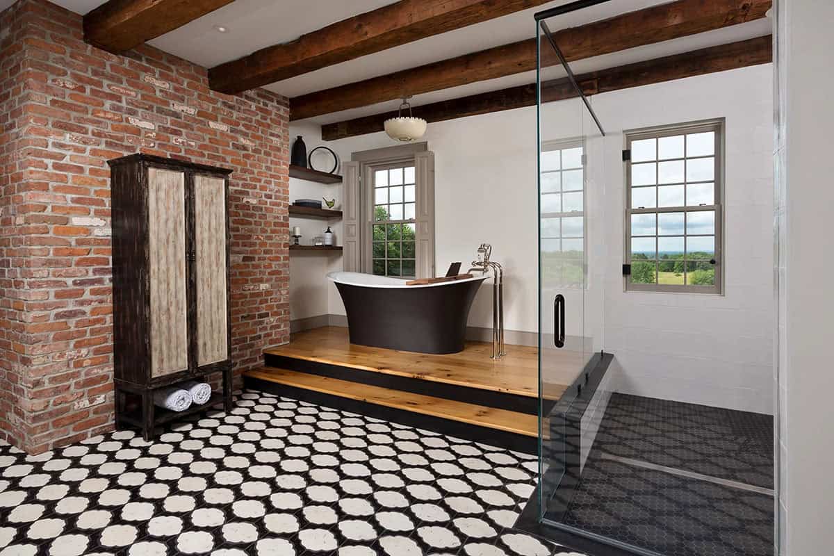 Primary bathroom with black freestanding soaking tub on raised wood platform, exposed brick wall, and patterned tile floor