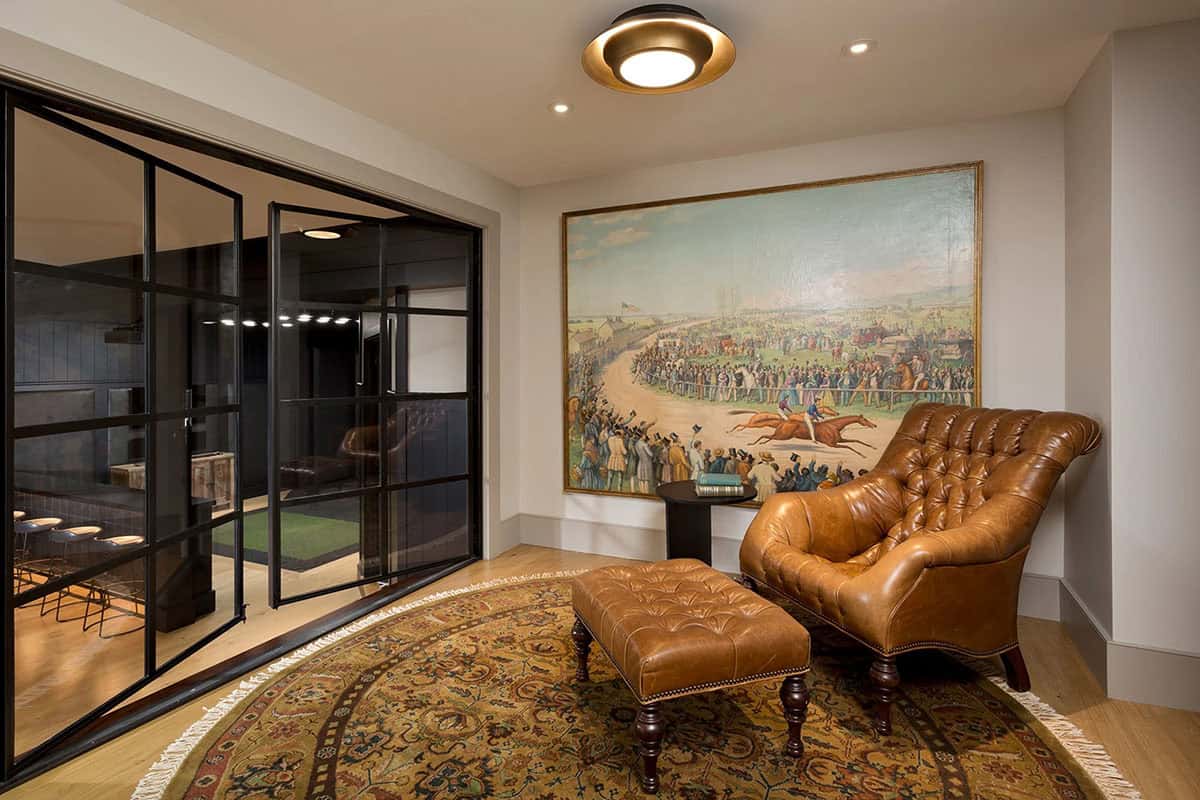 Reading nook with tufted leather chair and ottoman, large equestrian painting, and steel-framed glass doors to golf simulator