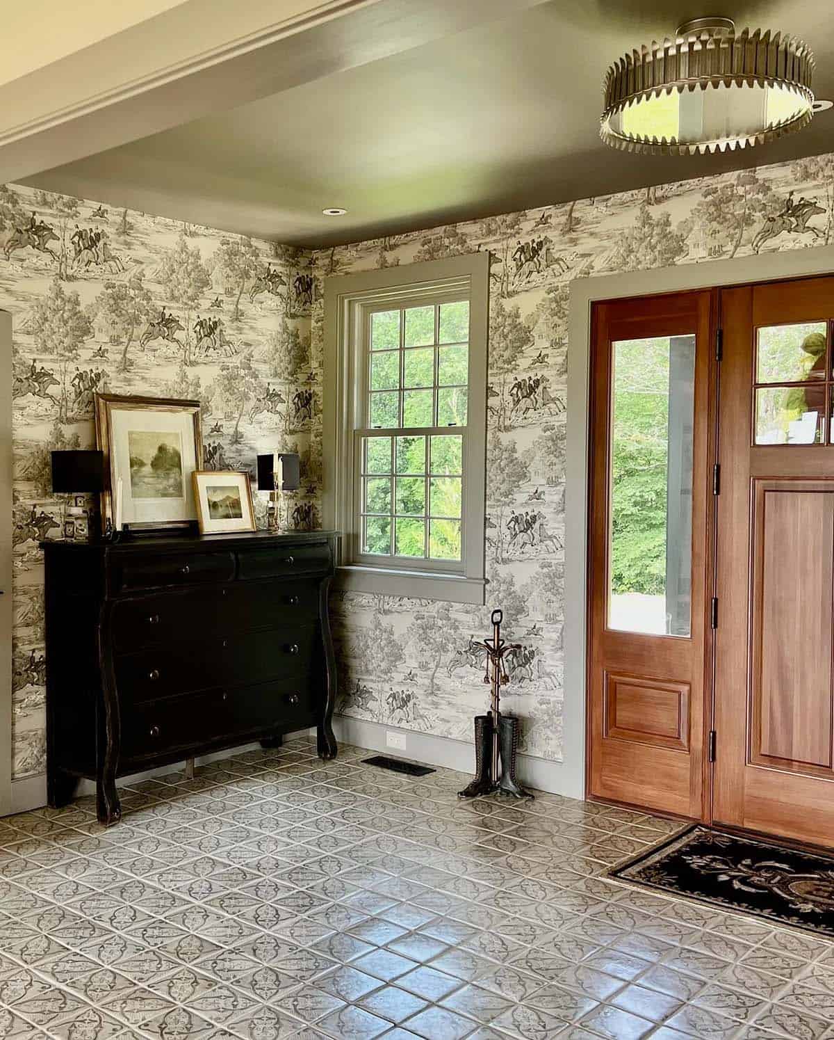 Farmhouse entry foyer with equestrian toile wallpaper, patterned cement tile floor, warm wood front door, and black dresser