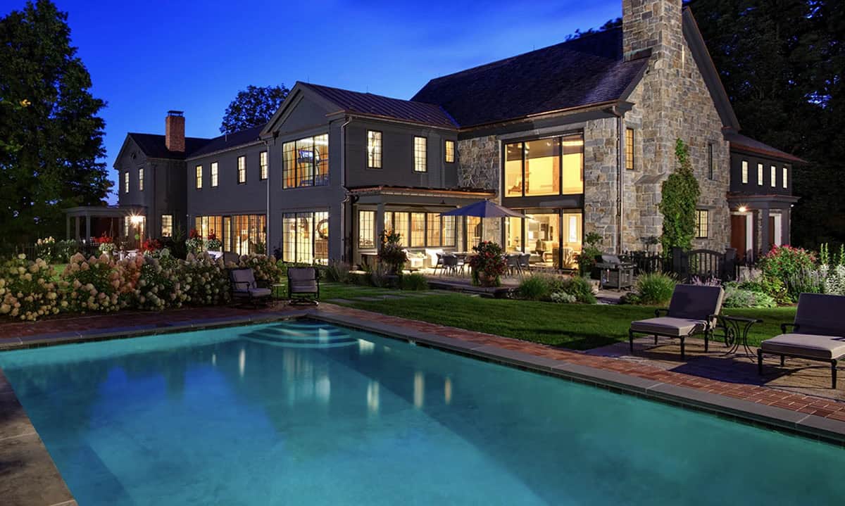 Illuminated rear facade of restored farmhouse at dusk with pool, brick surround, outdoor seating, and stone addition glowing warmly