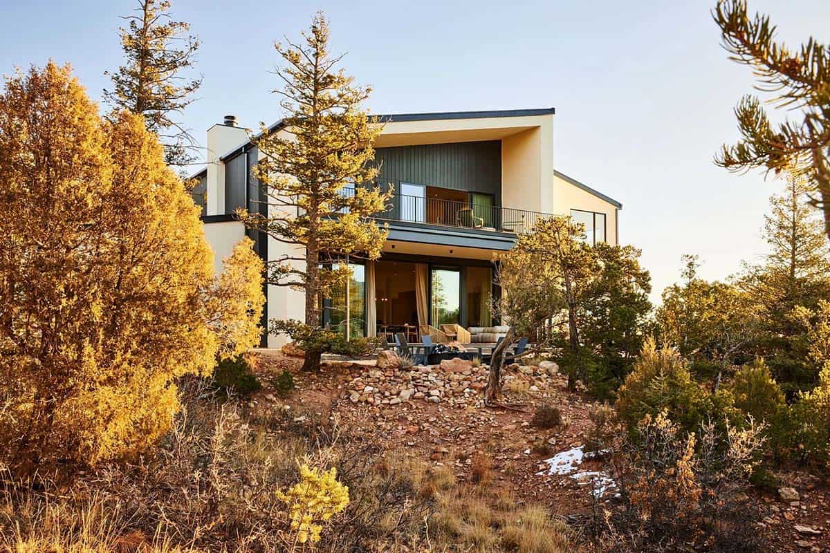 Modern two-story mountain home with flat roof, dark vertical siding, and outdoor patio seating amid golden autumn trees in Colorado