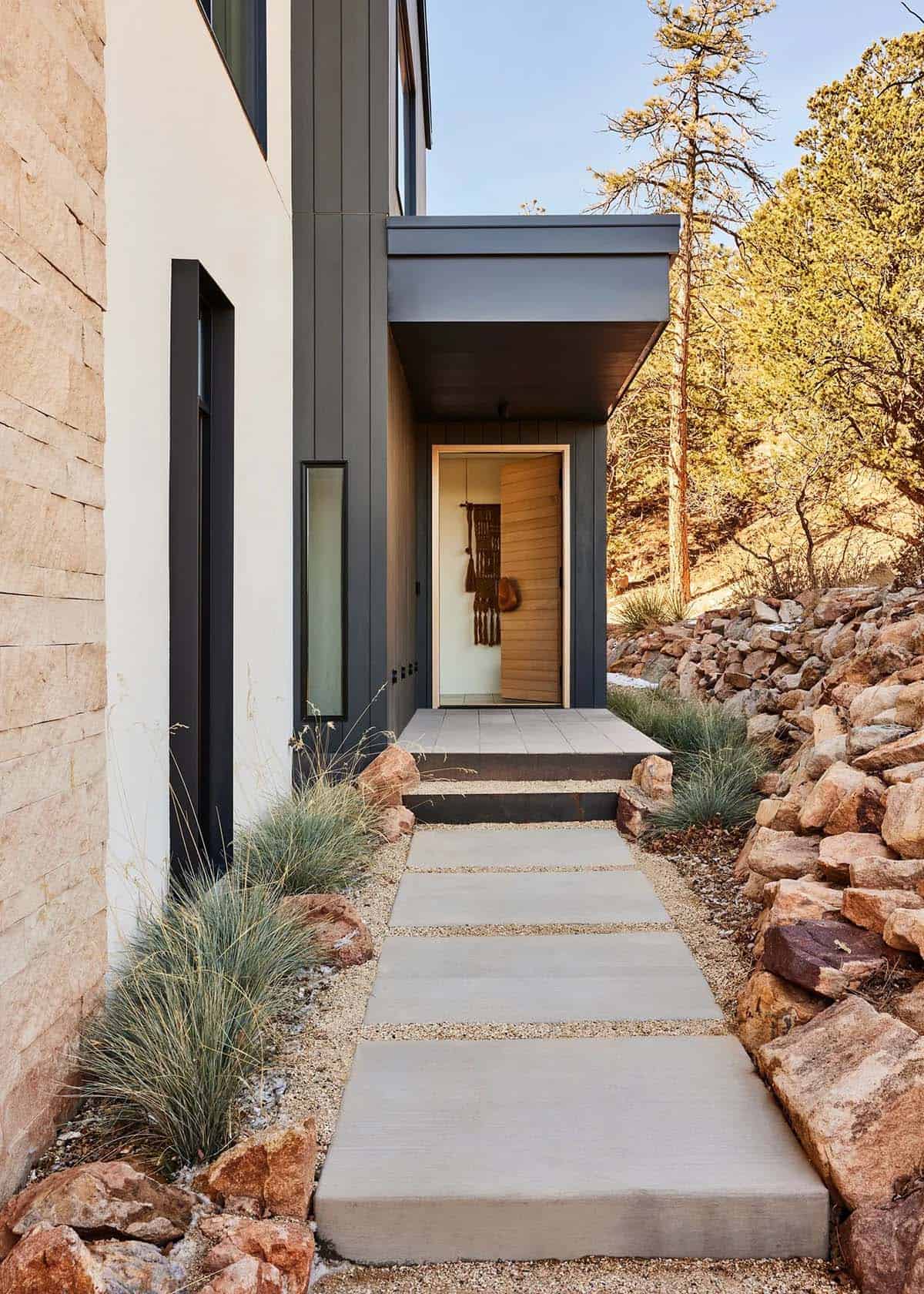 Contemporary home entry with concrete stepping stones, ornamental grasses, and a warm wood pivot door framed by dark steel