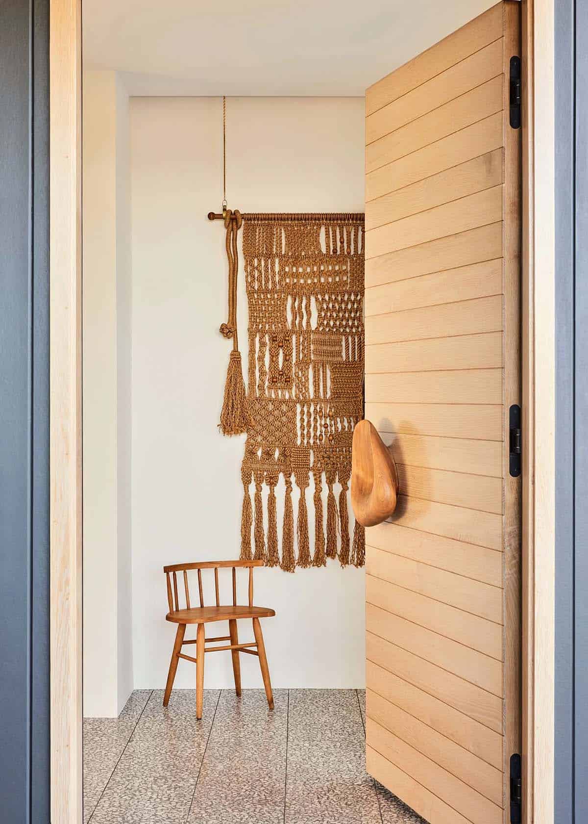 Entryway with a large woven macram&eacute; wall hanging, simple wood chair, and terrazzo tile floor beside a slatted wood door