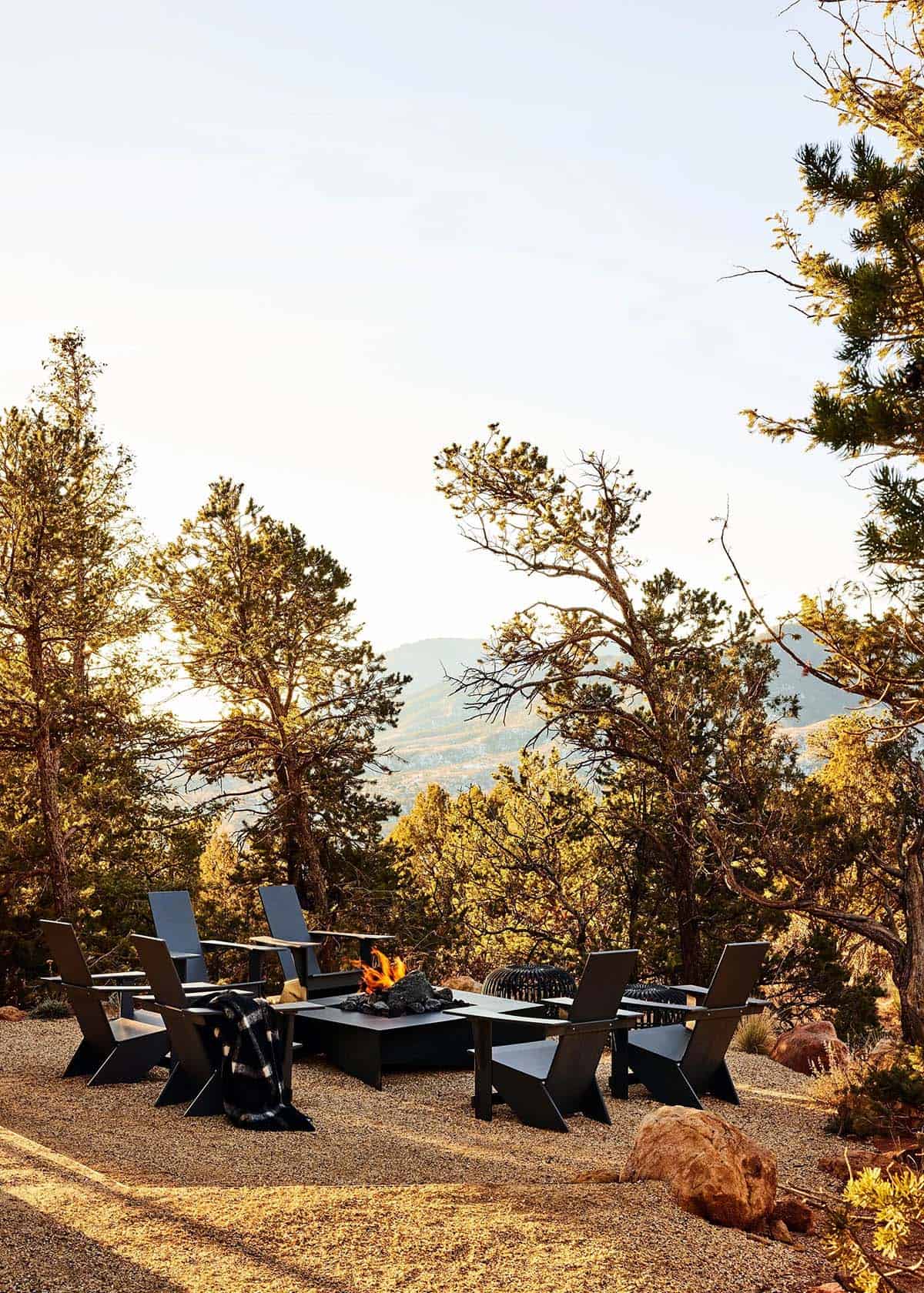 Outdoor fire pit seating area with black modern Adirondack chairs surrounded by pine trees and mountain views