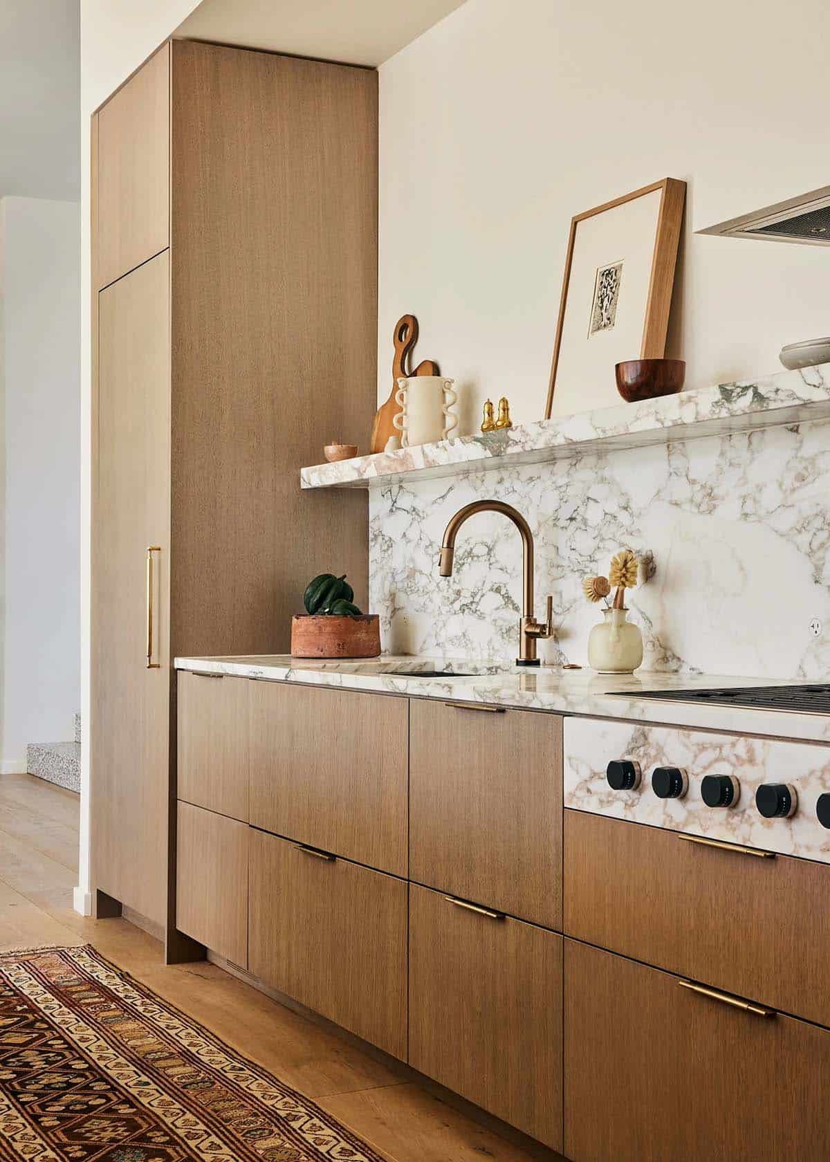 Kitchen wall with warm walnut cabinetry, marble countertop and backsplash, brass faucet, and Persian runner on hardwood floor