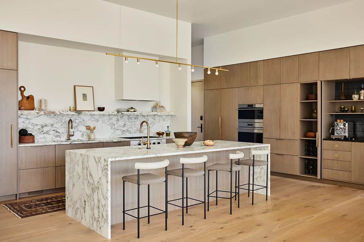 Open-plan kitchen with large marble waterfall island, four upholstered barstools, brass linear pendant light, and floor-to-ceiling oak cabinetry