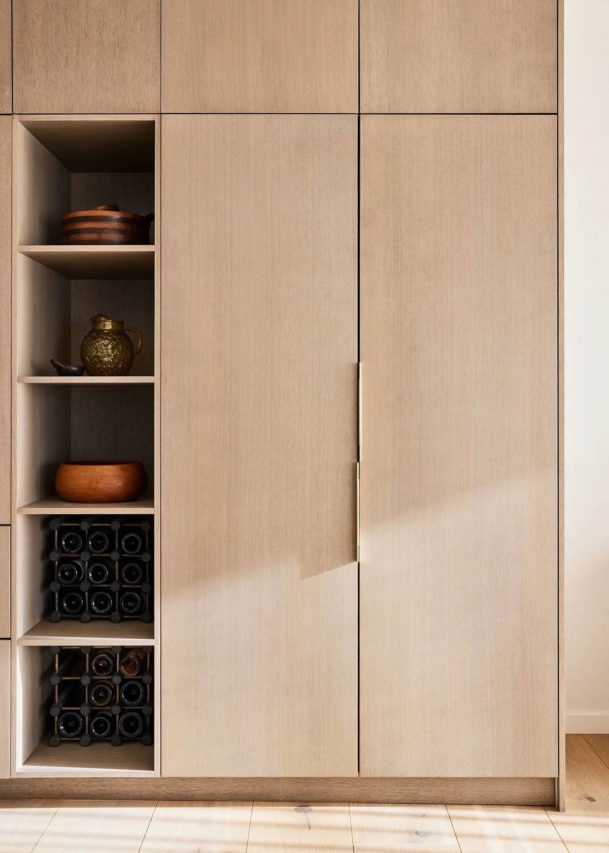 Floor-to-ceiling oak cabinetry with open shelving displaying ceramics and built-in wine storage, with a brass pull handle