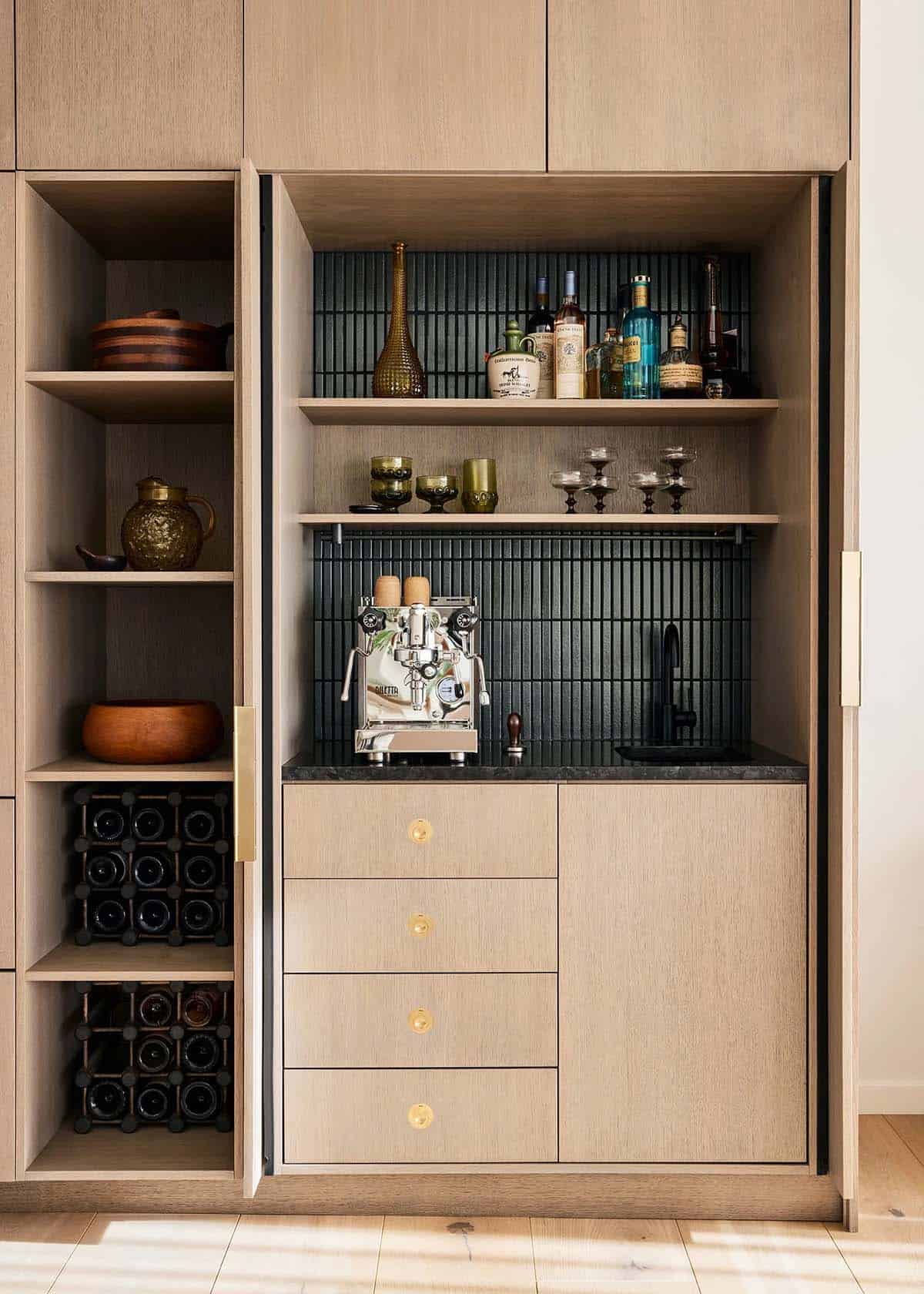 Built-in oak bar cabinet open to reveal dark tile backsplash, espresso machine, liquor bottles, glassware, and wine rack