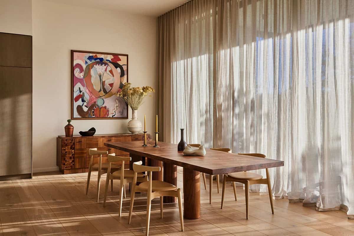 Dining room with dark walnut table, light oak chairs, sheer linen curtains, sculptural sideboard, and colorful abstract painting