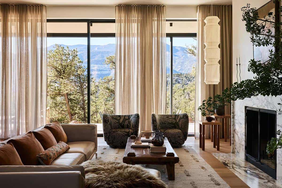 Living room with floor-to-ceiling windows framing mountain views, leather sofa, patterned armchairs, marble fireplace, and paper pendant lamp