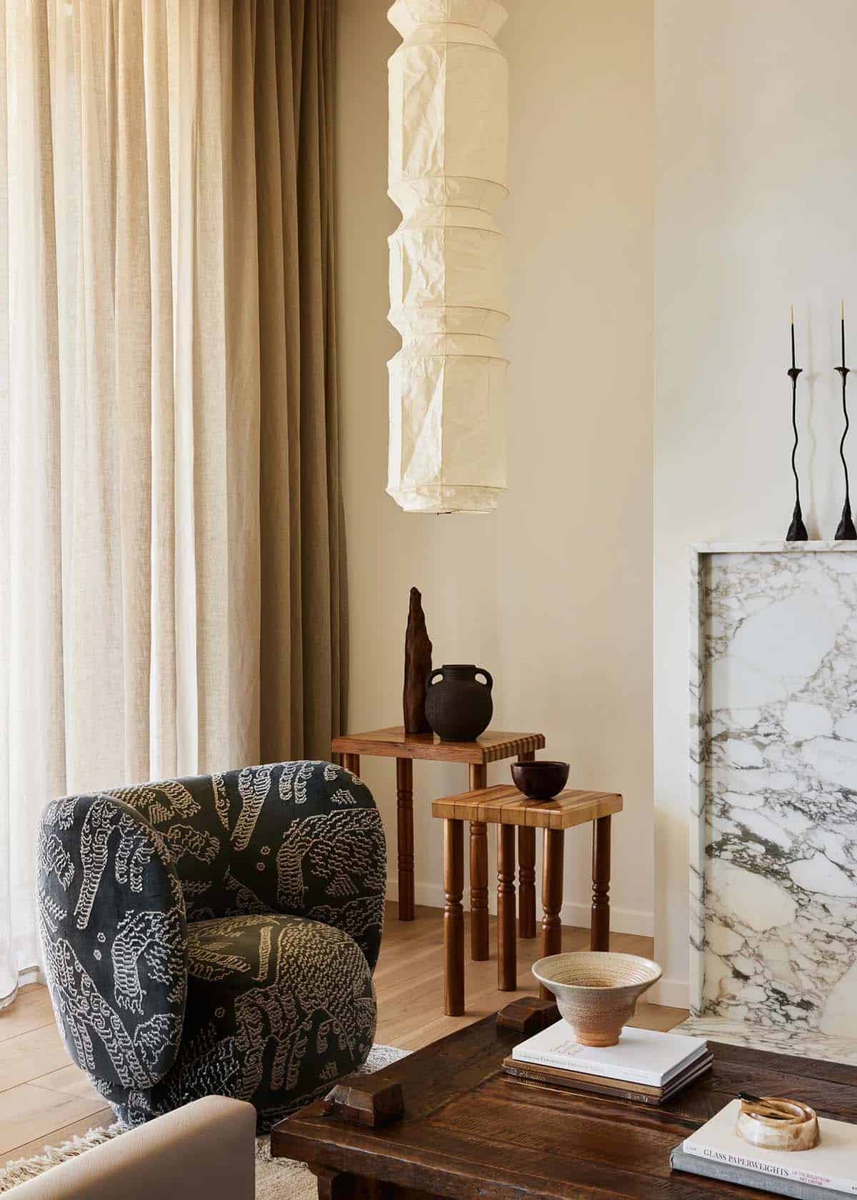 Living room vignette with patterned armchair, paper lantern pendant, marble fireplace surround, and layered wood side tables with ceramics